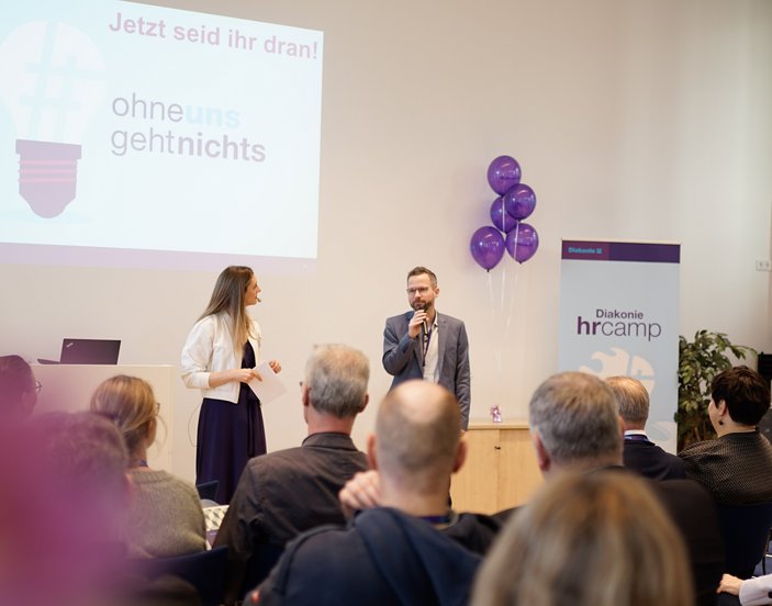 Eine Frau und ein Mann stehen mit Mikrofonen auf dem Podium einer Veranstaltung der Diakonie