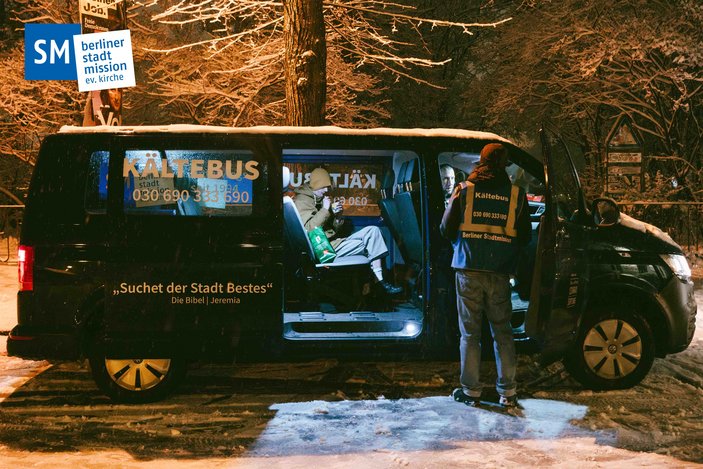 Zwei Menschen sitzen in einem offenen Bus, der bei Schneefall an der Straße parkt. 