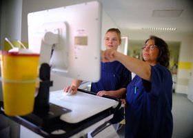 Zwei Pflegekräfte stehen vor einem Bildschirm und zeigen mit dem Finger auf den Monitor