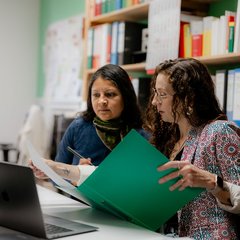 Zwei Frauen sitzen zu einem Beratungsgespräch am Schreibtisch vor einem Laptop, die eine Frau hält eine Aktenmappe in der Hand