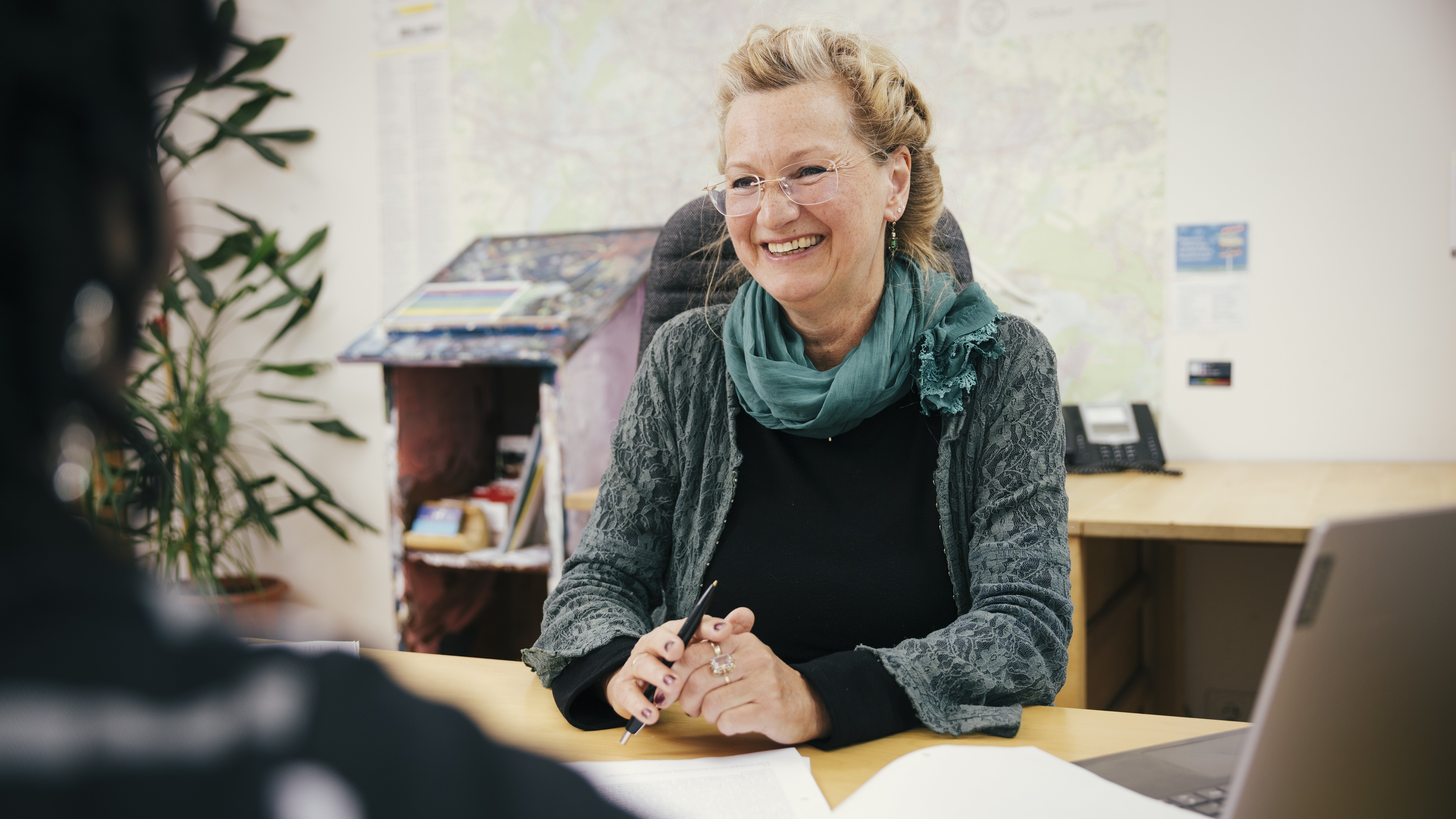 Frau lächelt, hält einen Stift in der Hand und hat Papiere auf dem Schreibtisch liegen