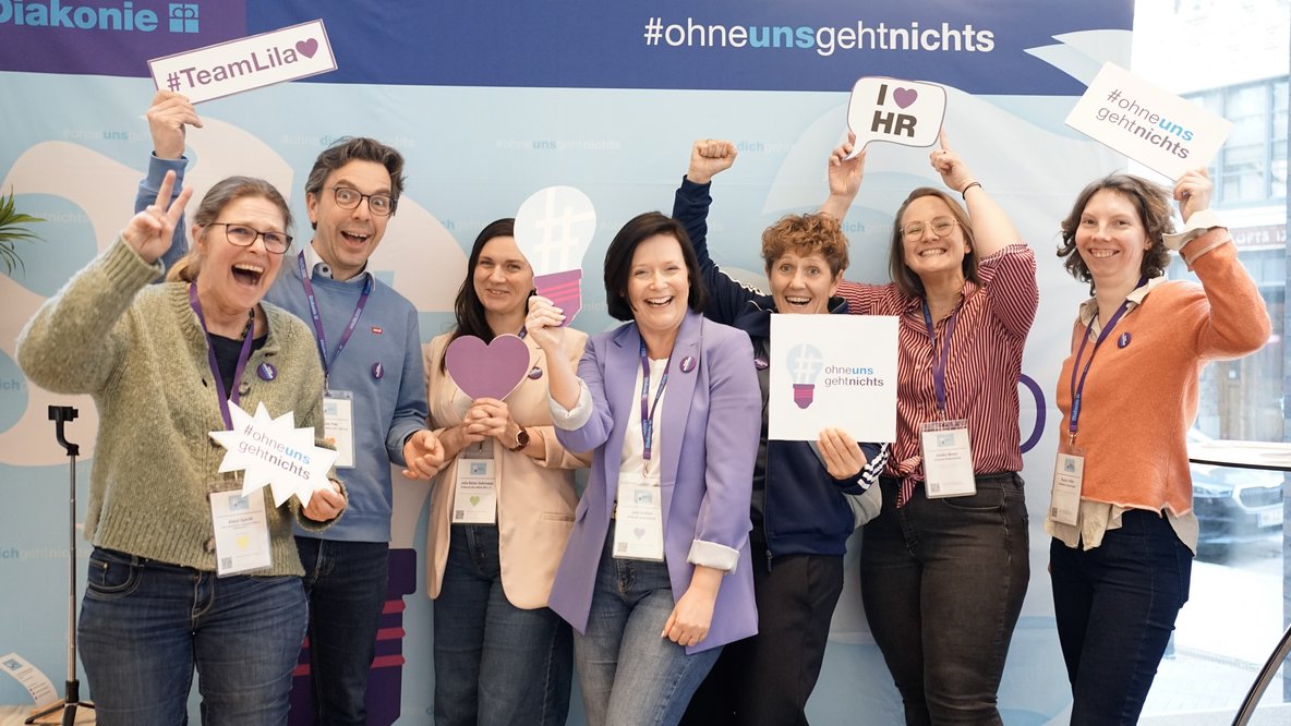 Das Organisationsteam des HR-Barcamps der Diakonie steht vor einer Stellwand und lacht in die Kamera