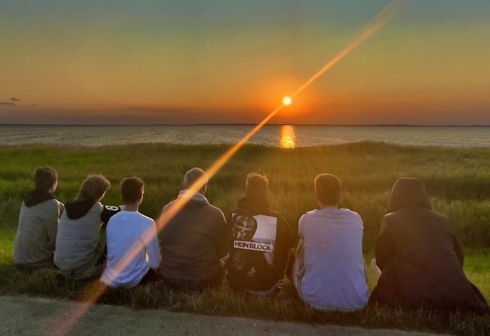 Man sieht eine Gruppe jugendlicher die am Ufer der Ostsee am Wegesrand sitzen und den Sonnenuntergang betrachten. Die rote Sonne steht schon nah über dem Horizont. Man kann das Ufer und das Wasser der Ostsee im Hintergrund erkennen. Durch die Aufnahme der Sonne entsteht ein Lichtstrahl der von links unten noch rechts oben durch die Sonne zu gehen scheint. Der Himmel hat einen Farbverlauf von oben blau nach unten rot.