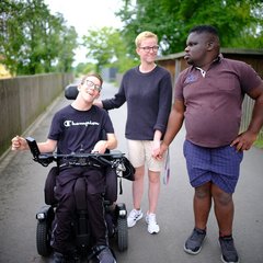 Eine Frau steht in der Mitte, links von ihr sitzt ein Mann im Rollstuhl, rechts von ihr hält sie einen Mann mit dunkler Hautfarbe an der Hand