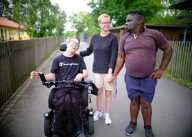 Eine Frau steht in der Mitte, links von ihr sitzt ein Mann im Rollstuhl, rechts von ihr hält sie einen Mann mit dunkler Hautfarbe an der Hand