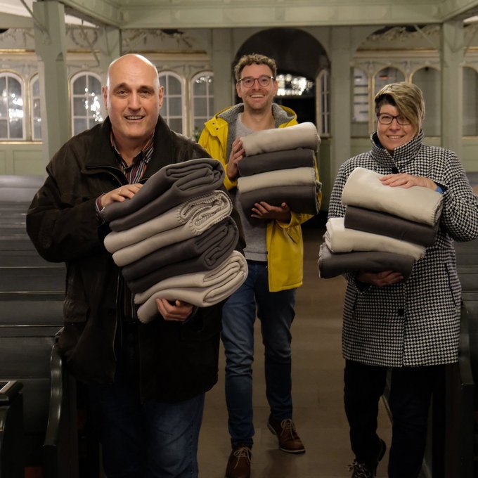 3 Menschen stehen mit Decken in einer Kirche für die Aktion wärmewinter
